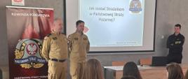 Na zdjęciu widać salę szkoleniową, w której odbywa się spotkanie informacyjne dla młodzieży.. Uczestnicy siedzą na krzesłach ustawionych w rzędach i słuchają prezentacji prowadzonej przez trzech funkcjonariuszy w umundurowaniu. Na środku znajduje się ekran z wyświetloną prezentacją dotyczącą służby w Państwowej Straży Pożarnej.