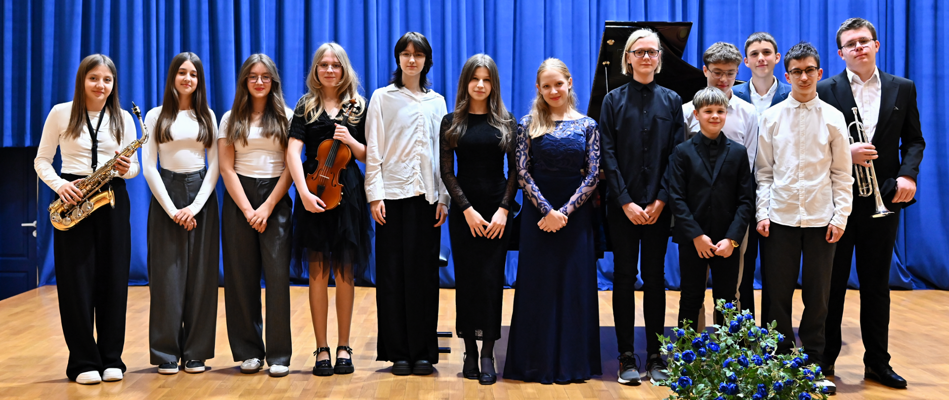 grupa kilkunastu dzieci na scenie w galowych strojach, niektórzy trzymają instrumenty muzyczne, za nimi niebieska kotara