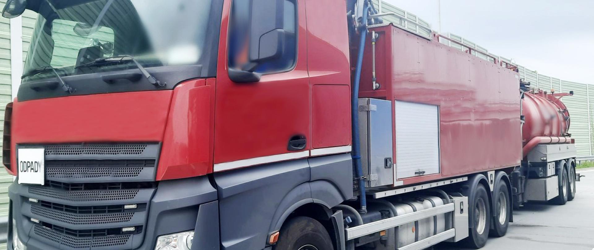 Zespół pojazdów zatrzymany przez inspektorów mazowieckiej Inspekcji Transportu Drogowego. Pojazd jest koloru czerwonego, z czarnymi i srebrnymi elementami, ma z przodu planszę z napisem ODPADY. Do pojazdu jest podpięta przyczepa, w podobnych kolorach. Zespół pojazdów stoi na przydrożnym parkingu. Za nim przydrożne ekrany akustyczne w kolorze zielonym.
