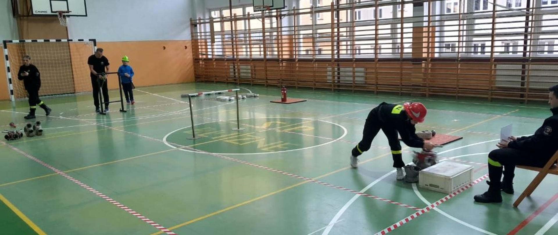 Kolorowa fotografia wykonana w sali gimnastycznej Szkoły Podstawowej w Bliznem. Przedstawia strażaka Młodzieżowej Drużyny Pożarniczej w czasie wykonywania zadania podczas zawodów halowych dla MDP. W tle sędziowie oraz inny zawodnik przygotowujący się do startu.