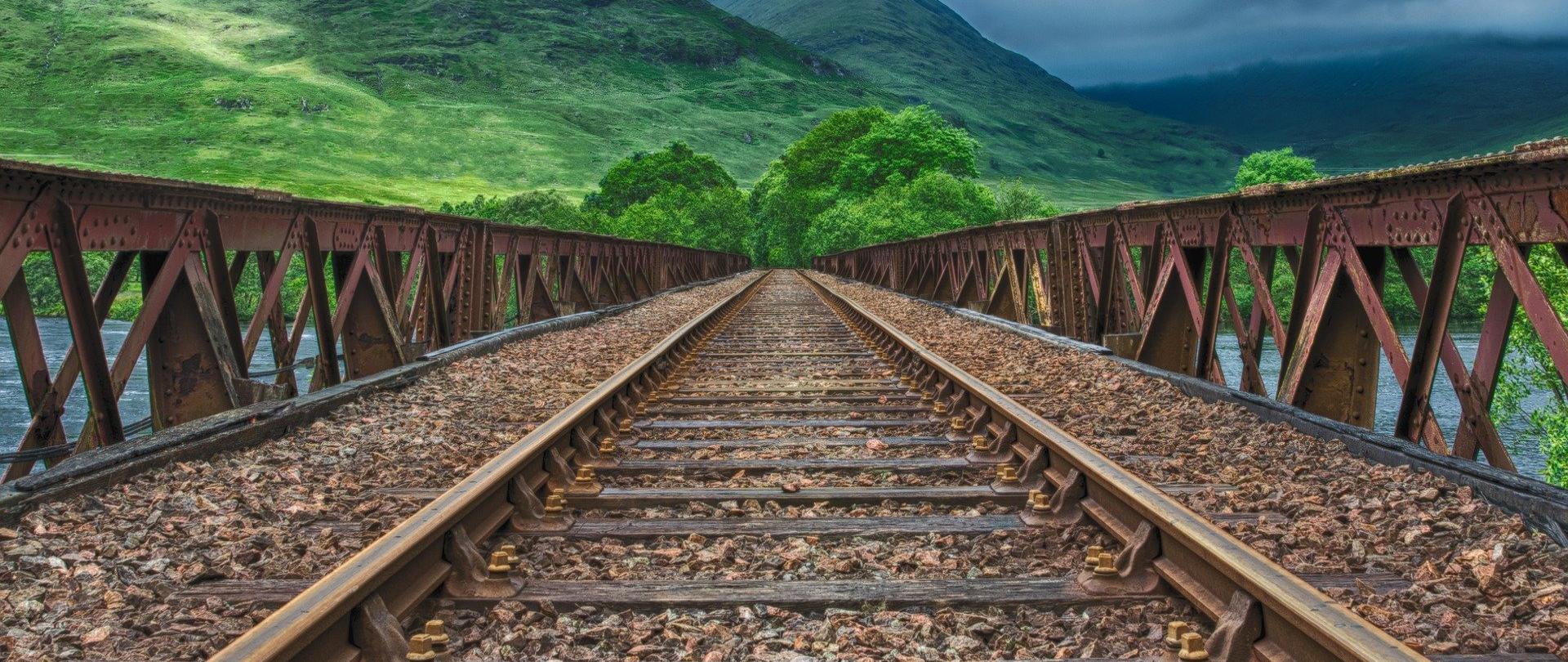 Zdjęcie przedstawia tory kolejowe prowadzące w dal przez wiadukt, w oddali szczyty gór.