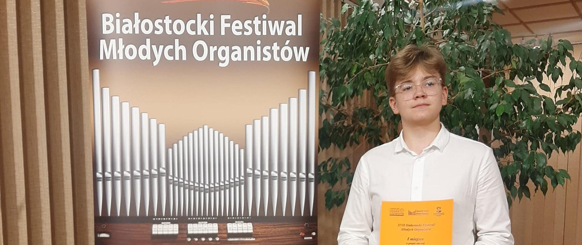 Po lewej stronie baner XVII Białostockiego Festiwalu Młodych Organistów, obok niego Bartosz Wesołek trzymający w rękach dyplom zajęcia I miejsca