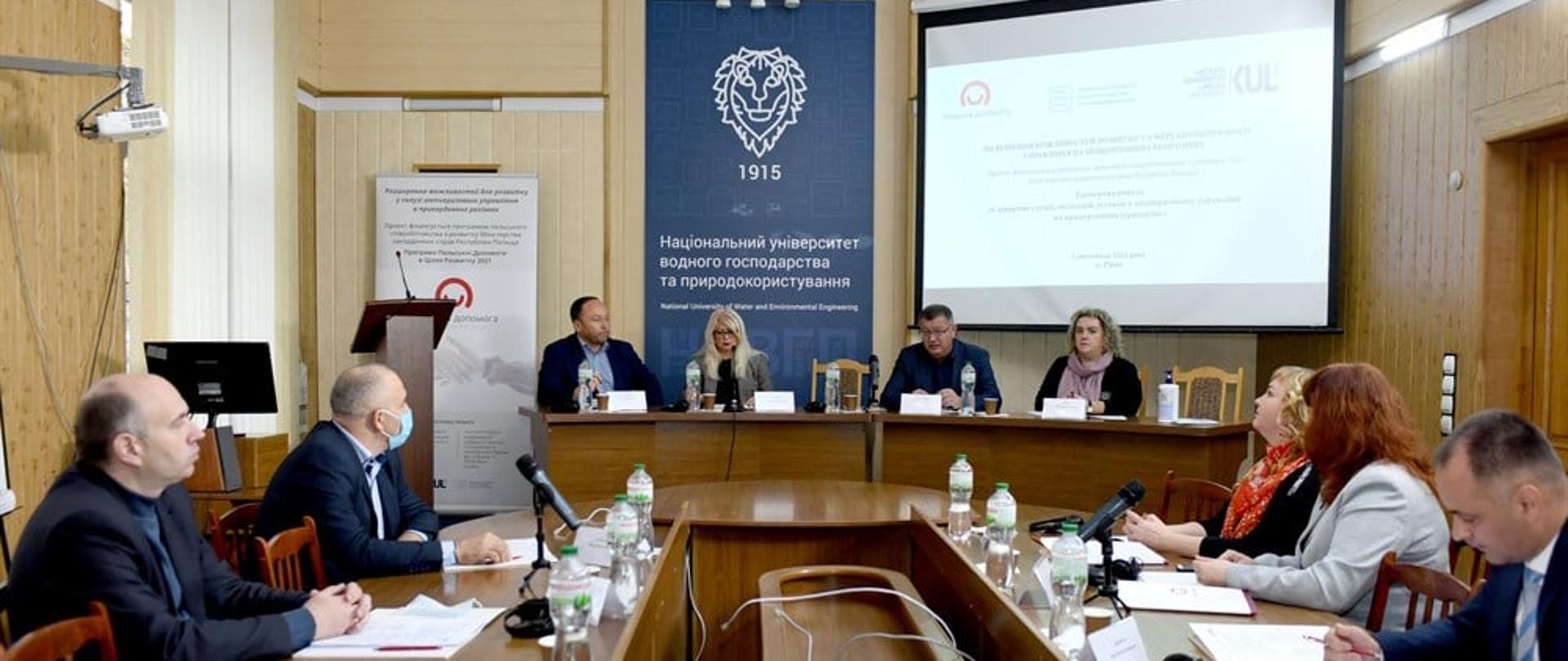 Grupa ludzi siedzi przy okrągłym stole i przy mównicy w eleganckim pomieszczeniu. 