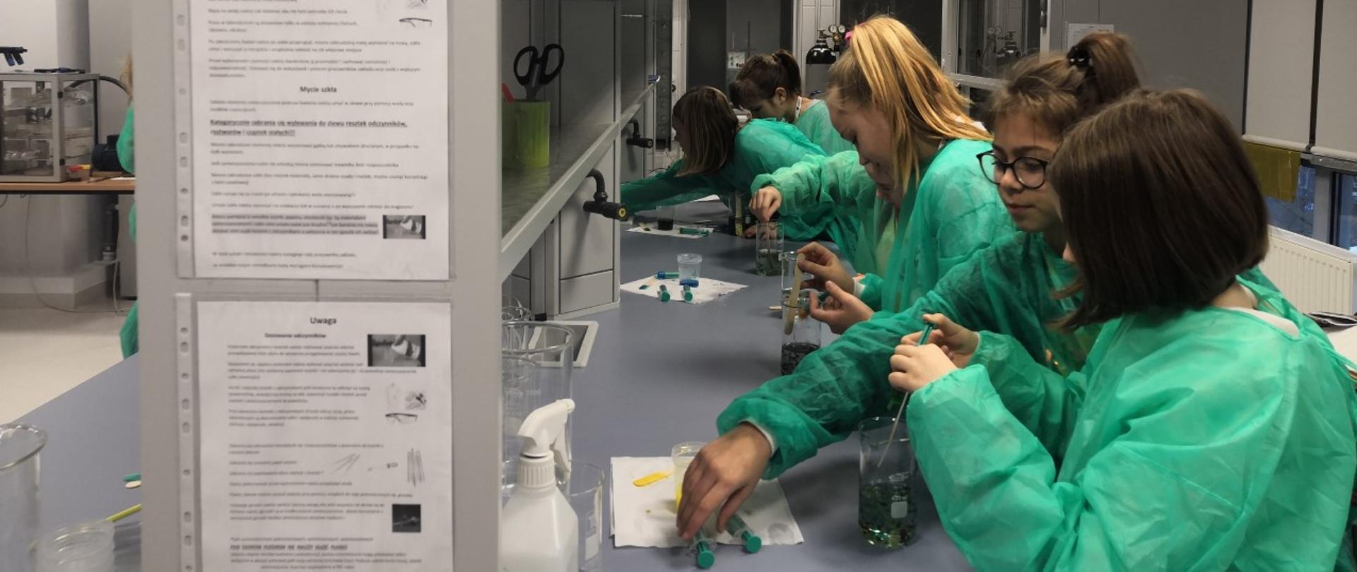 Zajęcia w laboratorium. Siedem nastolatek w zielonych fartuchach, stojąc w rzędzie przy długim blacie, wlewa do menzurek płyn za pomocą strzykawek