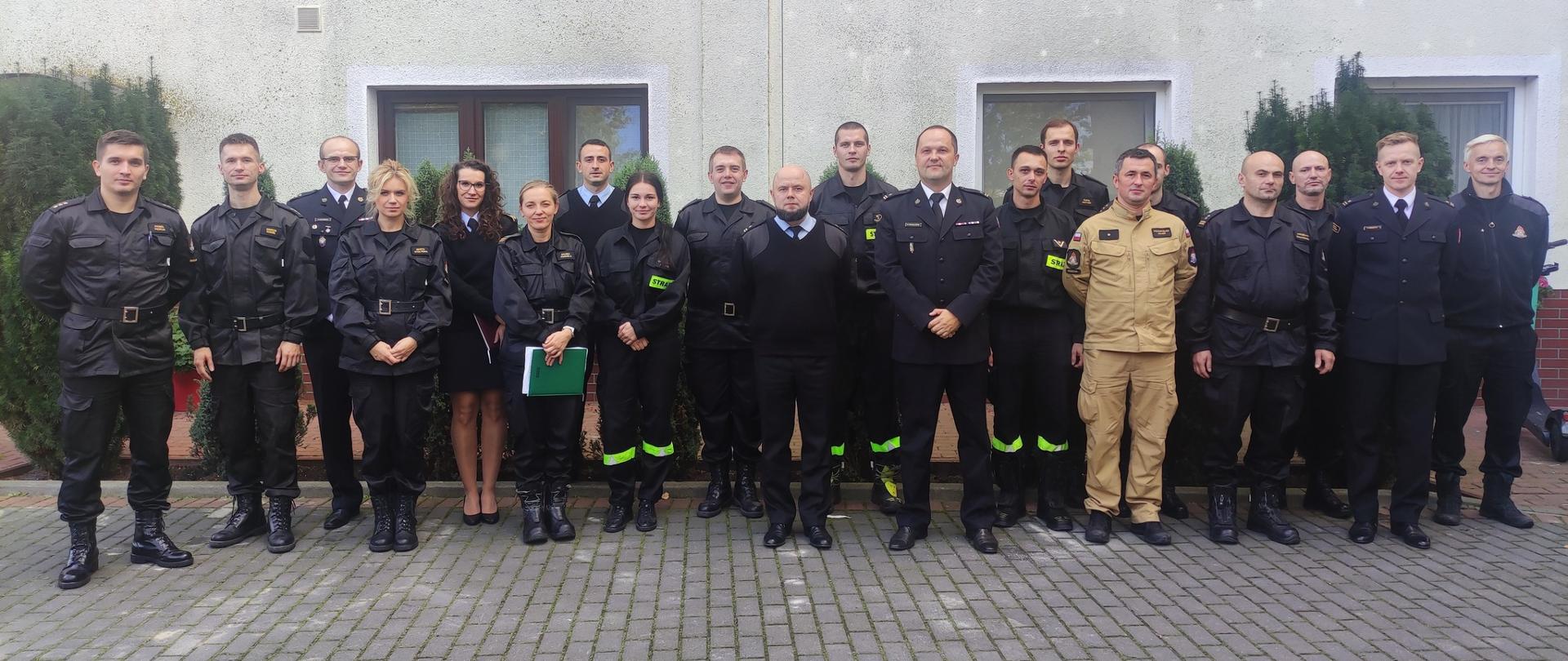 Na zdjęciu przed budynkiem stoi w dwuszeregu grupa kilkunastu osób w czarnym umundurowaniu i jeden mężczyzna w piaskowym.
