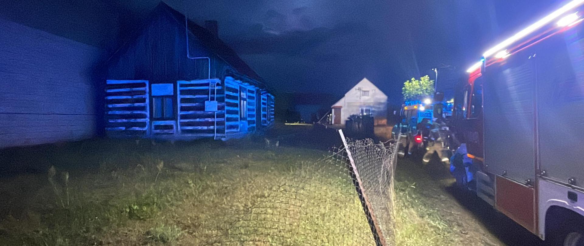Nocna scena przedstawia akcję ratunkową w budynku. Po lewej stronie widoczny jest długi, niski, drewniany budynek. W tle kolejny dom jest słabo oświetlony. Po prawej stronie kadru widoczna jest przednia część wozu strażackiego z migającymi czerwonymi i niebieskimi światłami. W pobliżu wozu widać kilku strażaków w odzieży ochronnej, a na pierwszym planie znajduje się druciane ogrodzenie. Teren porośnięty jest trawą. Cała scena sugeruje nocną akcję służb ratunkowych, prawdopodobnie na terenie wiejskim.
