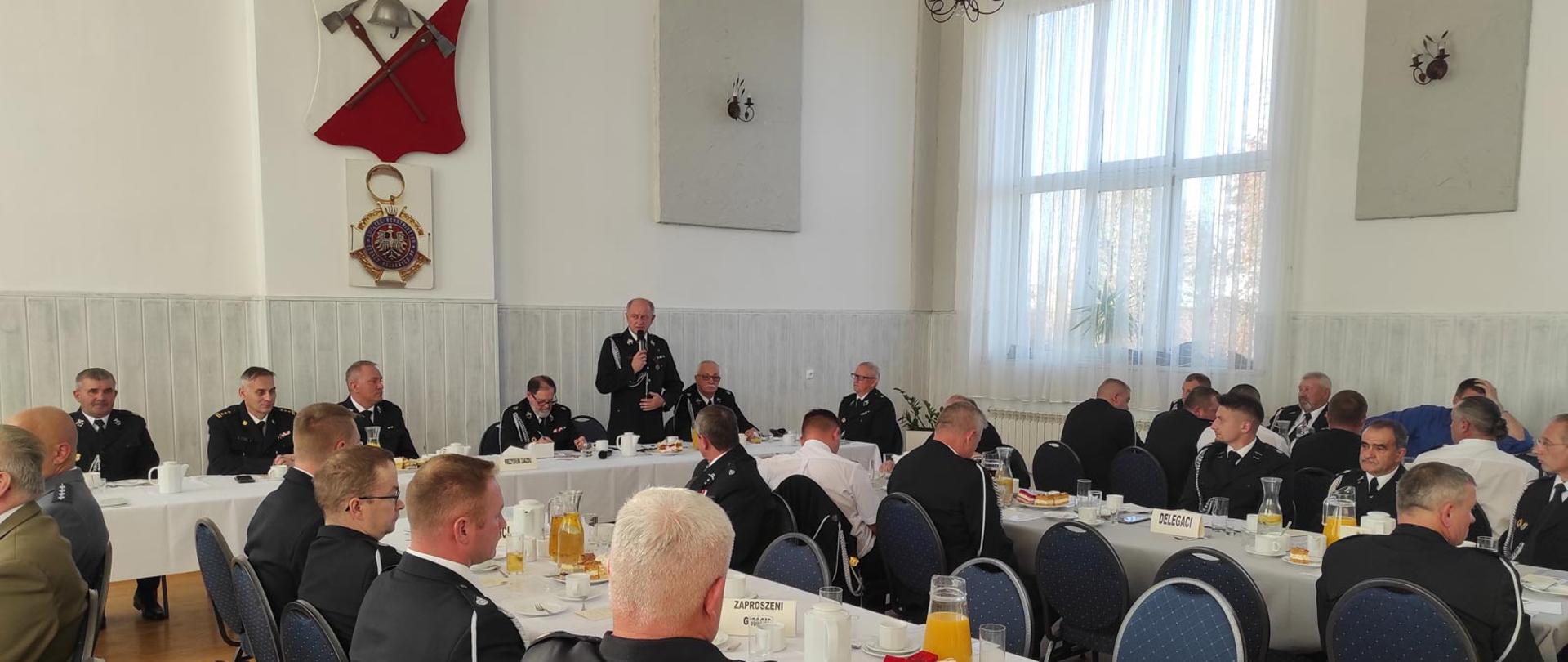 Zjazd Oddziału Gminnego Związku Ochotniczych Straży Pożarnej RP w Koszęcinie. Prezydium zjazdu siedzące za stołem oraz strażacy siedzący przy stołach.
