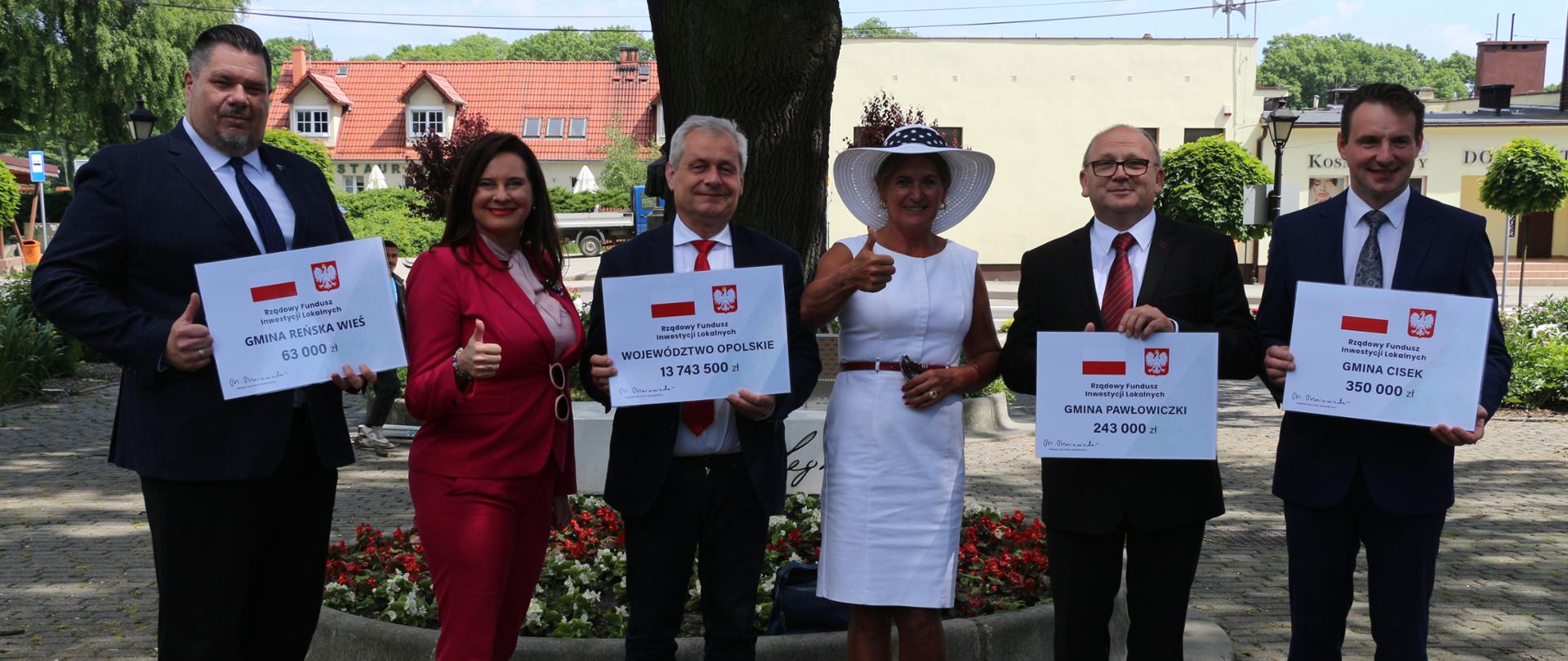 Na zdjęciu 6 osób, stojących obok siebie. Pozują do zdjęcia. Od lewej wójt gminy Reńska Wieś - postawny mężczyzna z brodą, trzymający w dłoni symboliczny czek. Nastepnie poseł Porowska, ubrana w czerwoną marynarkę, unosi kciuk do góry na znak zwycięstwa. Obok niej, w centralnej cześć kadru, wojewoda opolski, który także trzyma kolejny czek w dłoni. Po prawej stronie wojewody stoi poseł Czochara, unosząca kciuk do góry. Na głowie ma biały kapelusz. Obok poseł dwóch mężczyzn - wójtowie gmin, trzymający czek w dłoniach. W tle fragment ulicy oraz okazały dąb.