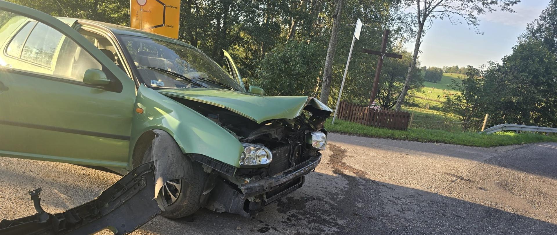 Pojazd osobowy marki VW koloru zielonego z uszkodzonym frontem pojazdu. W tle widoczny jest przydrożny krzyż oraz tereny zielone. Na jezdni widoczna jest plama płynów eksploatacyjnych.