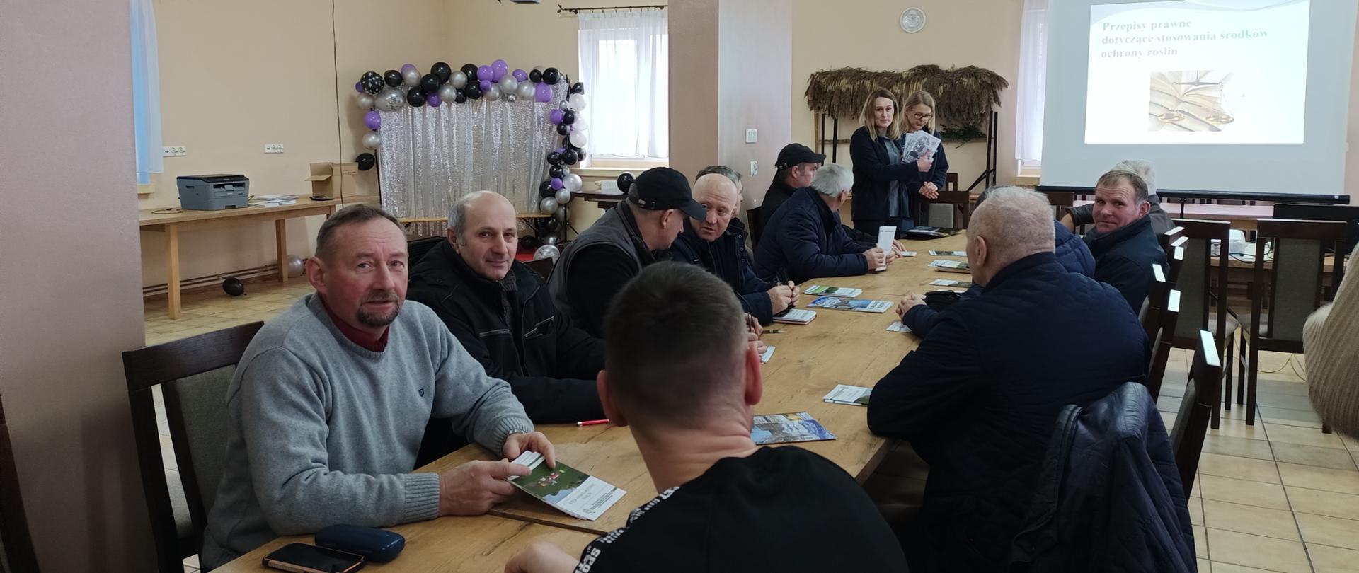  Uczestnicy siedzą po obu stronach stołu, w głębi zdjęcia stoją dwie osoby, jedna trzyma w ręku ulotki. w tle slajd z prezentacji przepisy prawne dotyczące stosowania środków ochrony roślin 