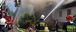 Zdjęcie ukazuje akcję gaśniczą w trakcie pożaru budynku pensjonatowego. Zdjęcie ukazuje strażaków ubranych w ubrania specjalne podających trzy prądy wody w natarciu na palący się budynek i jeden prąd wody w natarciu z podnośnika hydraulicznego . Kłęby dymu wydobywają się spod dachu.