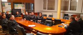 Na zdjęciu widać funkcjonariuszy KM PSP w Kaliszu. Ubrani są w umundurowanie dowódczo-sztabowe koloru czarnego. Osoby siedzą przed stołem. Zwróceni są w stronę przewodniczącego spotkania. Stoły koloru brązowego. Pomieszczenie oświetlone sztucznym światłem. Podłoga z paneli podłogowych.