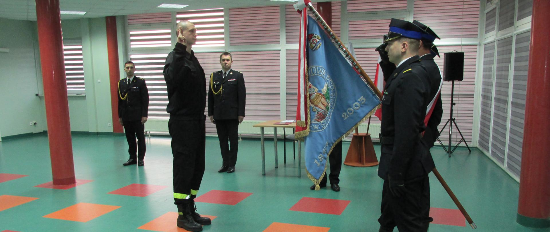 Na zdjęciu w centralnej jego części stoi w pozycji zasadniczej w umundurowaniu koszarowym strażak Bartłomiej Miśkowicz. Strażak podzas ślubowania bez nakrycia głowy z uniesioną prawą ręką. W tle za strażakiem po lewej stronie stoi w pozycji zasadniczej w umundurowaniu wyjściowym asp. Marcin Łojek natomiast z prawej stoi w pozycji zasadniczej w umundurowaniu wyjściowym Komendant Powiatowy Państwowej Straży Pożarnej w Strzyżowie brygadier Rafał Rutyna. Obok Komendanta widoczny stolik z dokumentami. W prawej części zdjęcia widoczny strażak odwrócony lewym bokiem w umundurowaniu wyjściowym który wchodzi w skład pocztu sztandarowego. Strażak oddaje honor przez salutowanie. Za strażakiem z przodu widoczny Sztandar Komendy Powiatowej Państwowej Straży Pożarnej w Strzyżowie. Sztandar opuszczony pod kątem około 45 stopni. Sztandar odwrócony błękitną stroną płata a na nim widoczne logo Państwowej Straży Pożarnej część haftu przedstawiającego Świętego Floriana oraz napis 1869 i 2003. Pod głowicą przymocowane wstęgi w barwach narodowych. Za strażakiem widoczna dolna część drzewca sztandaru. Dalej w tle czarny głośnik na statywie.