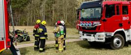 Grupa strażaków w pełnym umundurowaniu i kaskach stoi na trawie obok czerwonego wozu strażackiego. Rozmawiają lub przygotowują się do działania. W tle widać las oraz sprzęt ratowniczy, w tym quad.