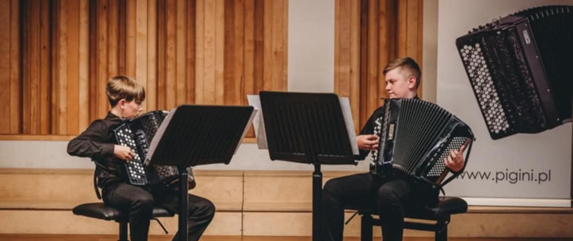 Na zdjęciu dwóch chłopców siedzi na krzesłach, grając na akordeonach. Obaj są ubrani w ciemne koszule i spodnie. Przed nimi znajdują się pulpity na nuty. Tło stanowią pionowe drewniane panele, a po prawej stronie widoczny jest pionowy baner z logo „PIGINI” i wizerunkiem akordeonu.