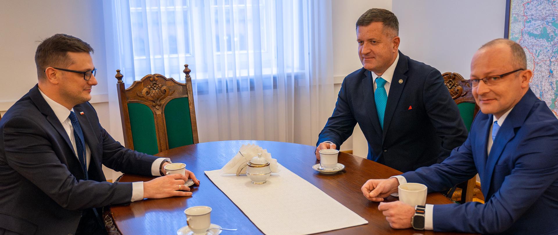 Na zdjęciu znajduje się trzech mężczyzn, którzy siedzą przy stole trzymając swoje filiżanki. Wiceminister Cyfryzacji Paweł Olszewski, Szef Służby Kontrwywiadu Wojskowego gen. bryg. dr Jarosław Stróżyk oraz Wojewoda Kujawsko-Pomorski Michał Sztybel. Po prawej stronie jest widoczny kawałek mapy Polski. 