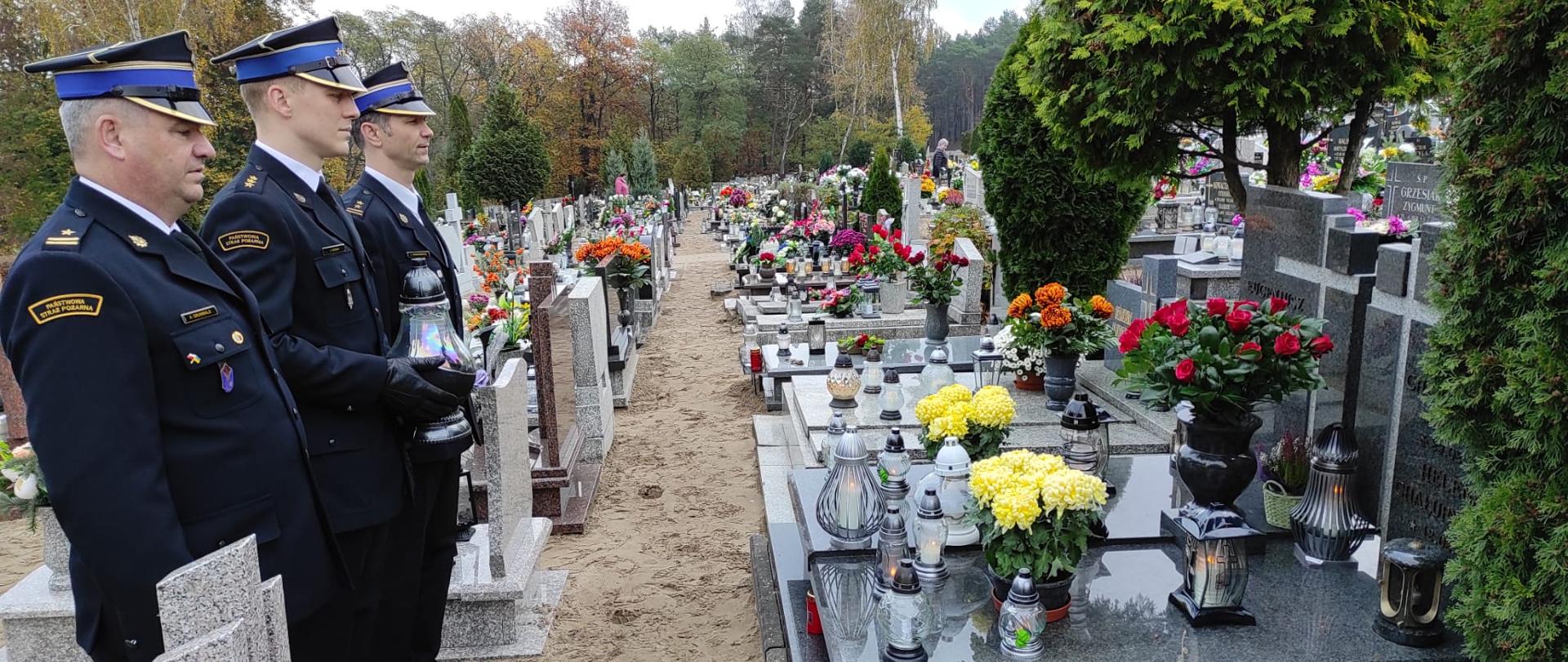 Zdjęcie przedstawia strażaków na cmentarzu, którzy salutują przy grobie.
W tle nagrobki i drzewa.
