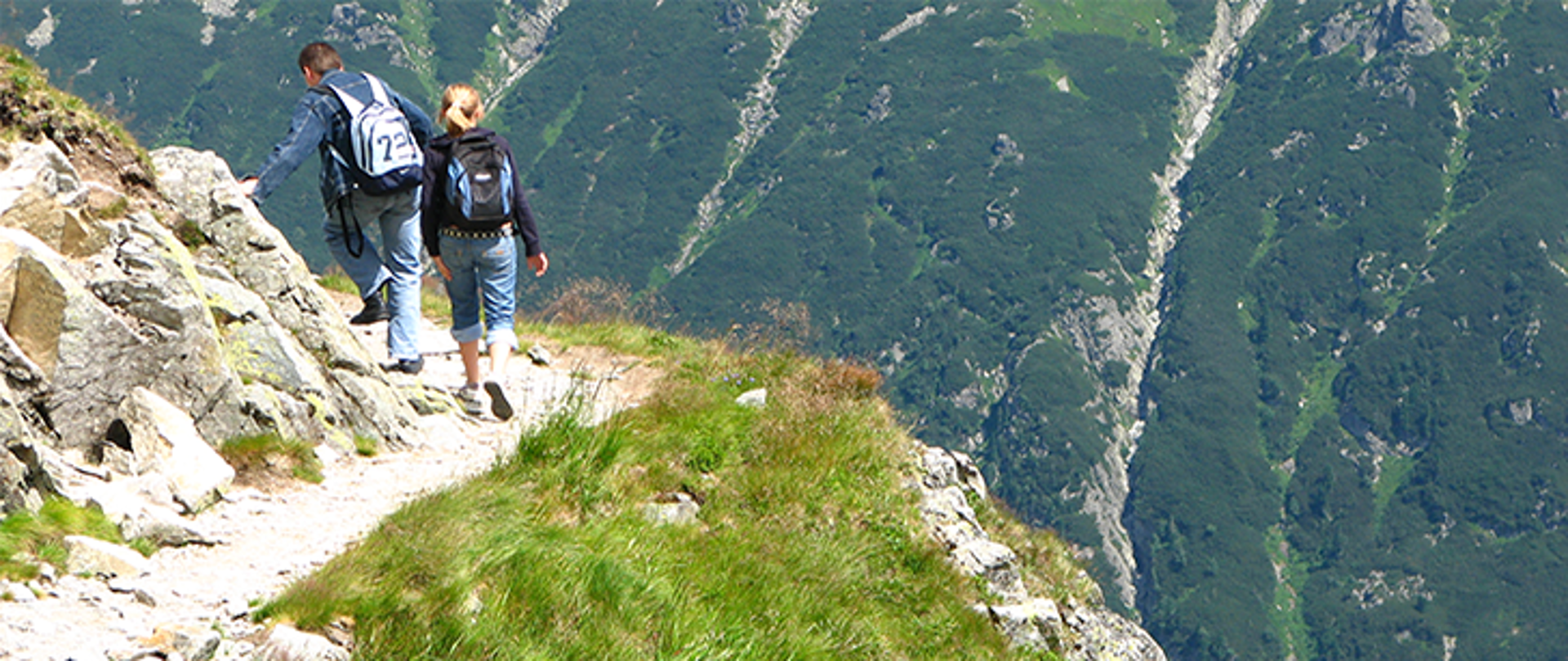 Zdjęcie. Po lewj stronie męższczyzna idziewczynka idą górską ścieżką. Po prawej stronie stromy stok.