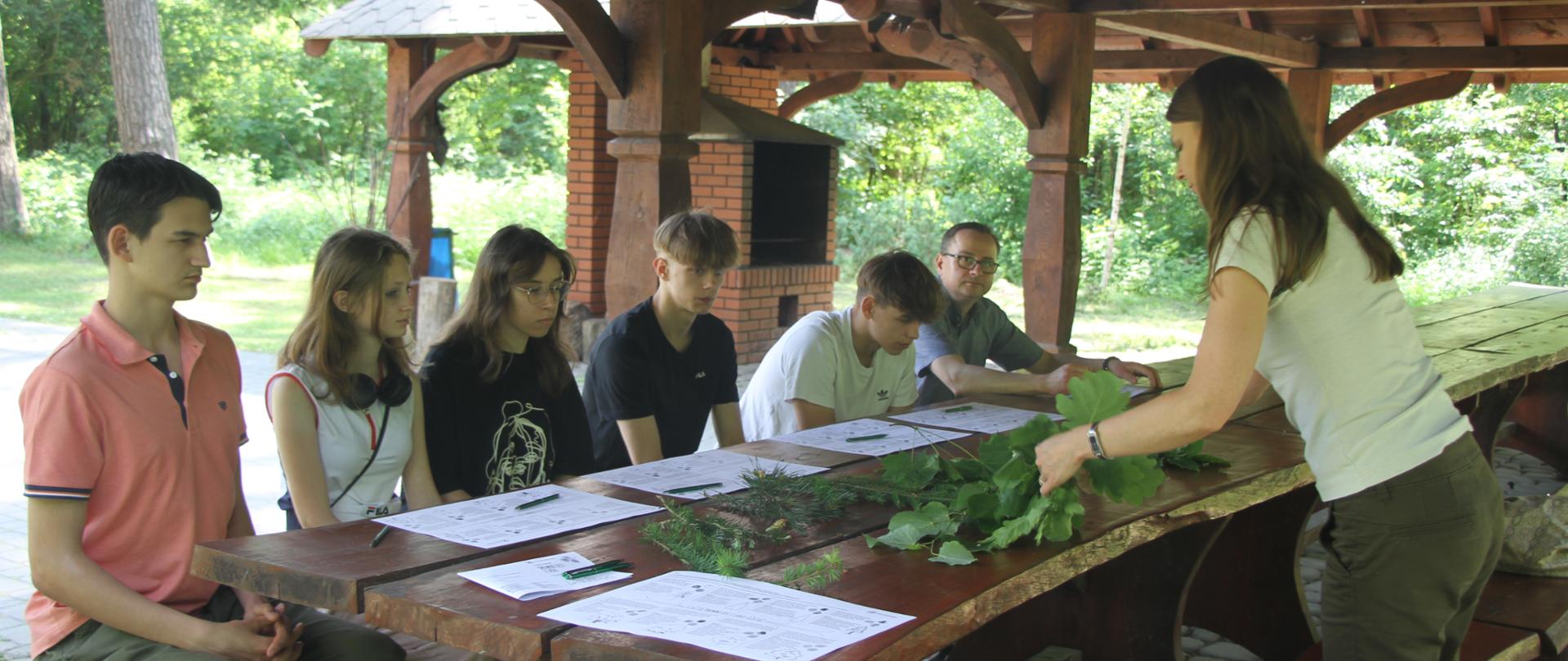 kilkuosobowa grupa siedzi przy dużym stole, na wprost nich kobieta prowadząca zajęcia i prezentująca różne liście drzew