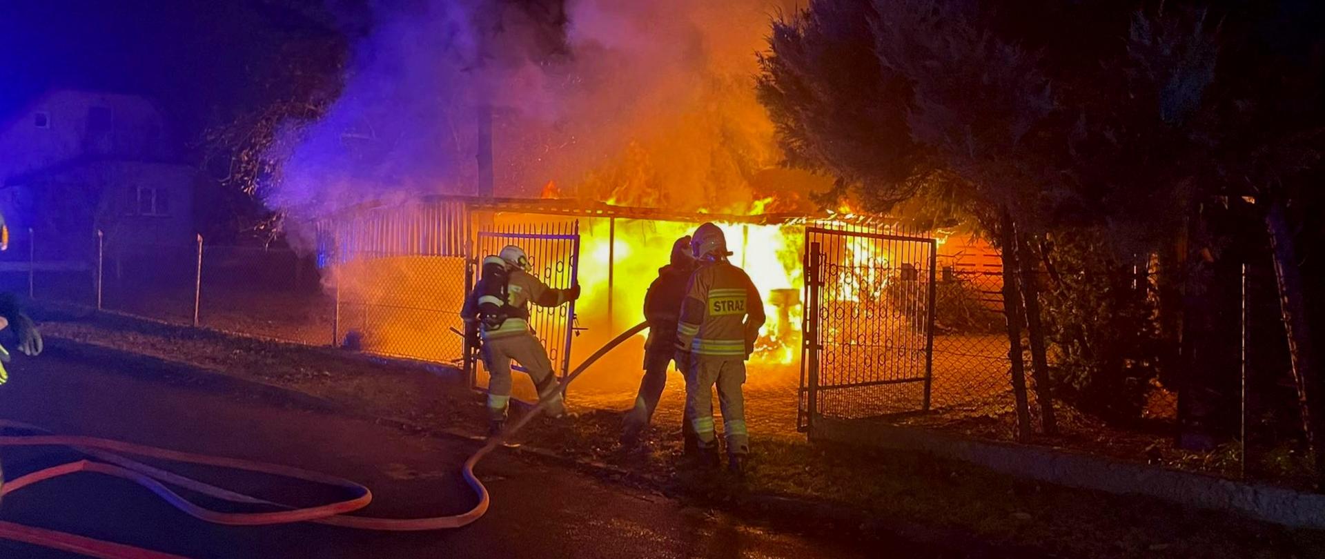 Zdjęcie przedstawia wiatę objętą ogniem. Przed wiatą dwóch strażaków: jeden otwiera bramę drugi trzyma wąż. Obok nich strażak dowódca. Na jezdni rozciągnięte węże. 