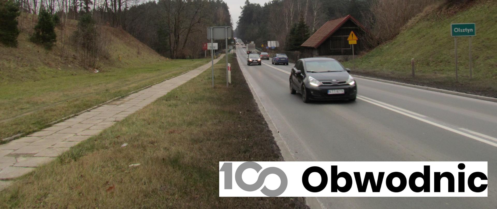 Asfaltowa droga wiodąca przez las. Po prawej stronie drogi tablica z nazwą miejscowości: Olsztyn.