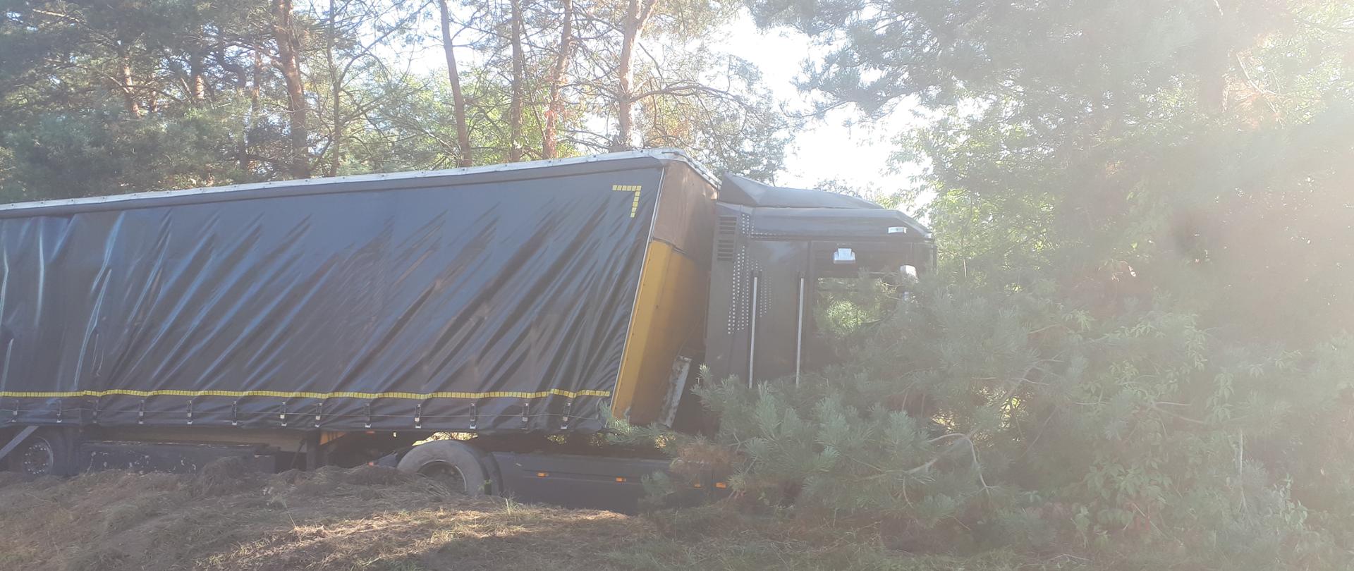 Zdjęcie przedstawia ciągnik siodłowy z naczepą, który zjechał z jezdni i stoi na poboczu zakopany w miękkim gruncie.