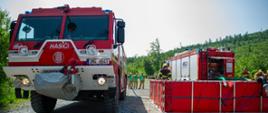 Czeski samochód gaśniczy Tatra z napisem „HASIČI” oraz polski wóz strażacki ustawione są obok zbiorników mobilnych typu open-top do czerpania wody. Zbiorniki są napełniane w ramach ćwiczeń z zaopatrzenia wodnego. W tle obserwatorzy i las.