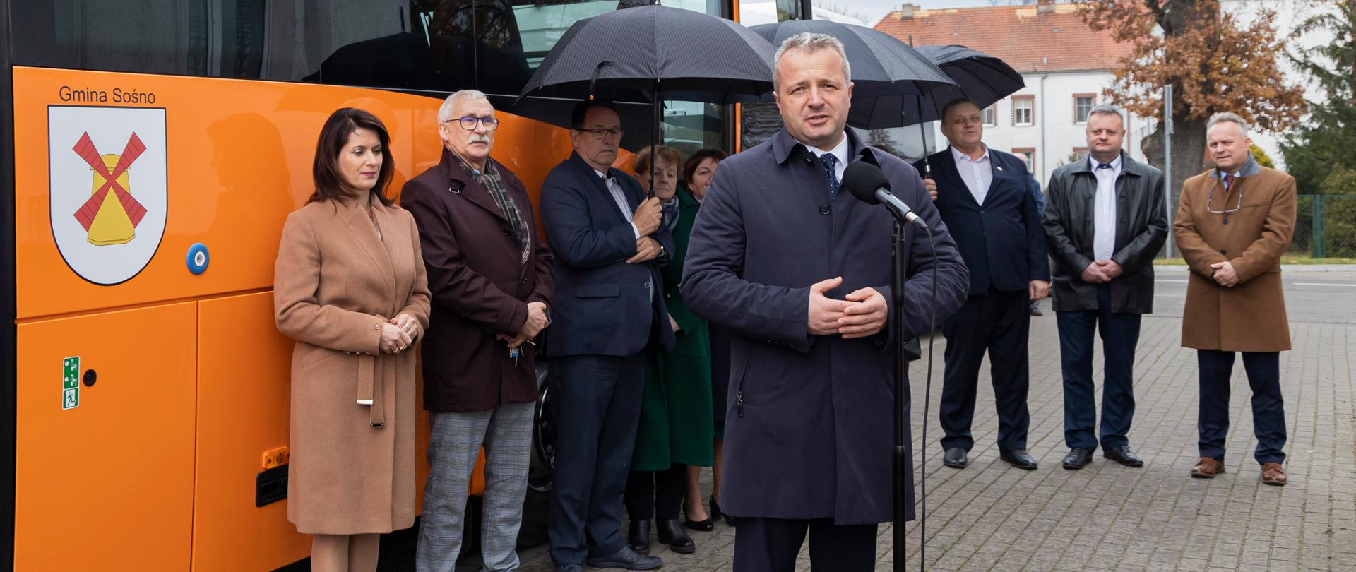 Jeden mężczyzna stojący przy mikrofonie i wypowiadający się. Za nim kilka osób