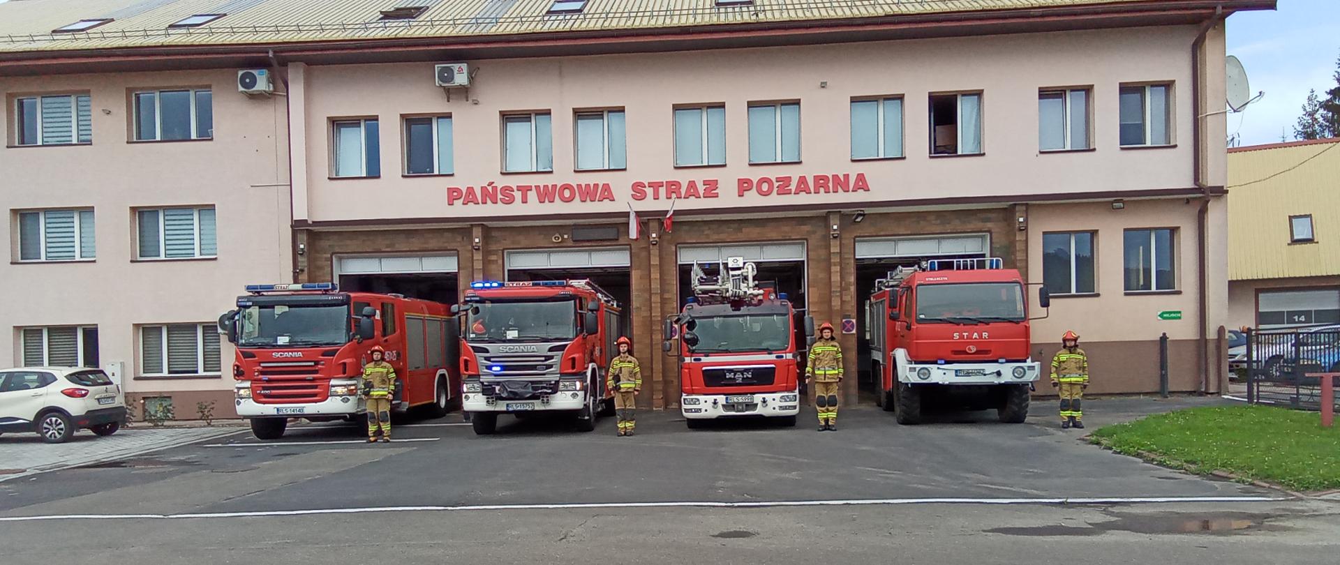 Zdjęcie przedstawia samochody ratowniczo – gaśnicze Jednostki ratowniczo-Gaśniczej Komendy Powiatowej Państwowej Straży Pożarnej w Lesku wraz z strażakami na tle budynku komendy. 