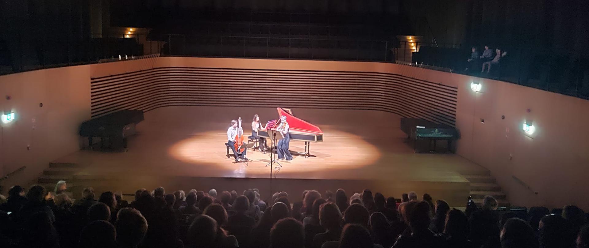 Dwie kobiety grają na skrzypcach i klawesynie, obok po lewej stronie mężczyzna siedzi i gra na violi da gamba na środku estrady sali koncertowej PSM , z przodu widać od tyłu zapełnioną publicznością widownię.
