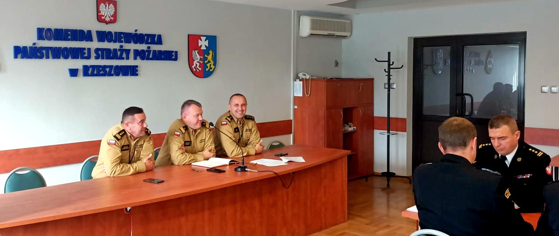 Zdjęcie zrobione w sali szkoleniowej. Przy stołach siedzą oficerowie Państwowej Straży Pożarnej uczestniczący w naradzie. Strażacy ubrani są mundury służbowe i wyjściowe.