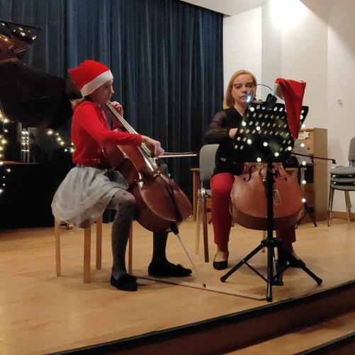 Na świątecznie przystrojonej scenie znajdują się uczennica i nauczycielka, grające na wiolonczelach.