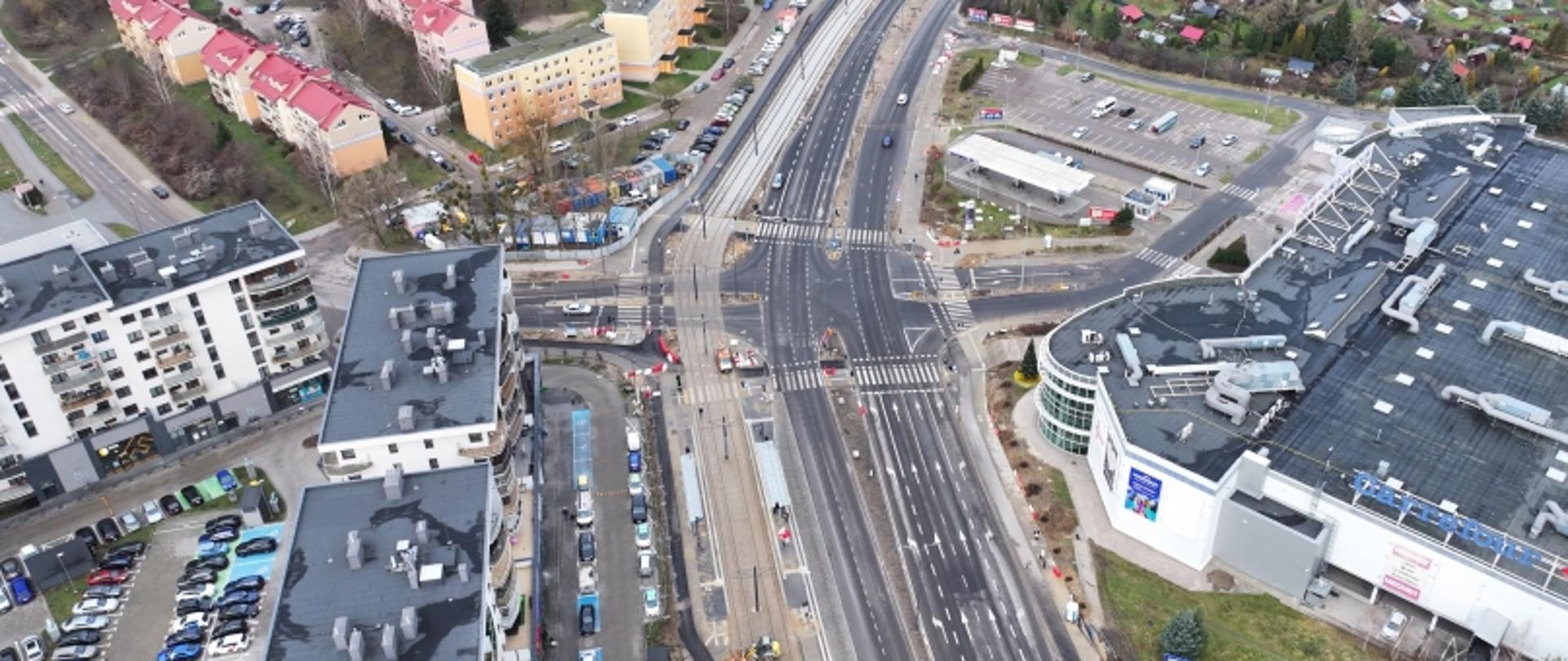 Zdjęcie lotnicze dużego skrzyżowania drogowego w Olsztynie, otoczonego budynkami mieszkalnymi i komercyjnymi. Widoczne nowe oznakowanie poziome, pasy ruchu i infrastruktura drogowa.