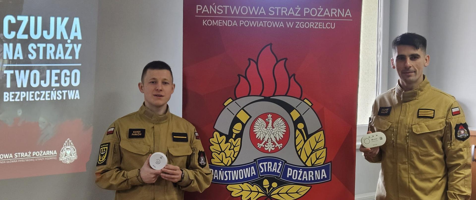 Na tle baneru informacyjnego KP PSP w Zgorzelcu pozuje do zdjęcia dwóch funkcjonariusz PSP ubranych w ubranie służbowe w kolorze piaskowym. Obydwaj funkcjonariusze trzymając w ręce czujki tlenku węgla.