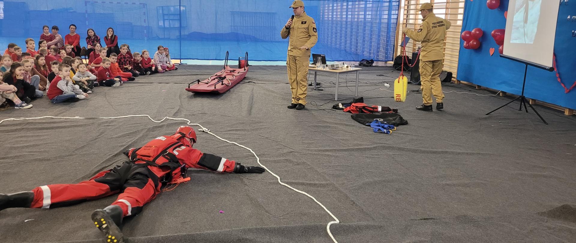 Jasna sala gimnastyczna, w której strażacy prowadzą zajęcia dla siedzących na podłodze dzieci i młodzieży z prezentacją sprzętu ratowniczego i z wykorzystaniem prezentacji multimedialnej wyświetlanej na ekranie.