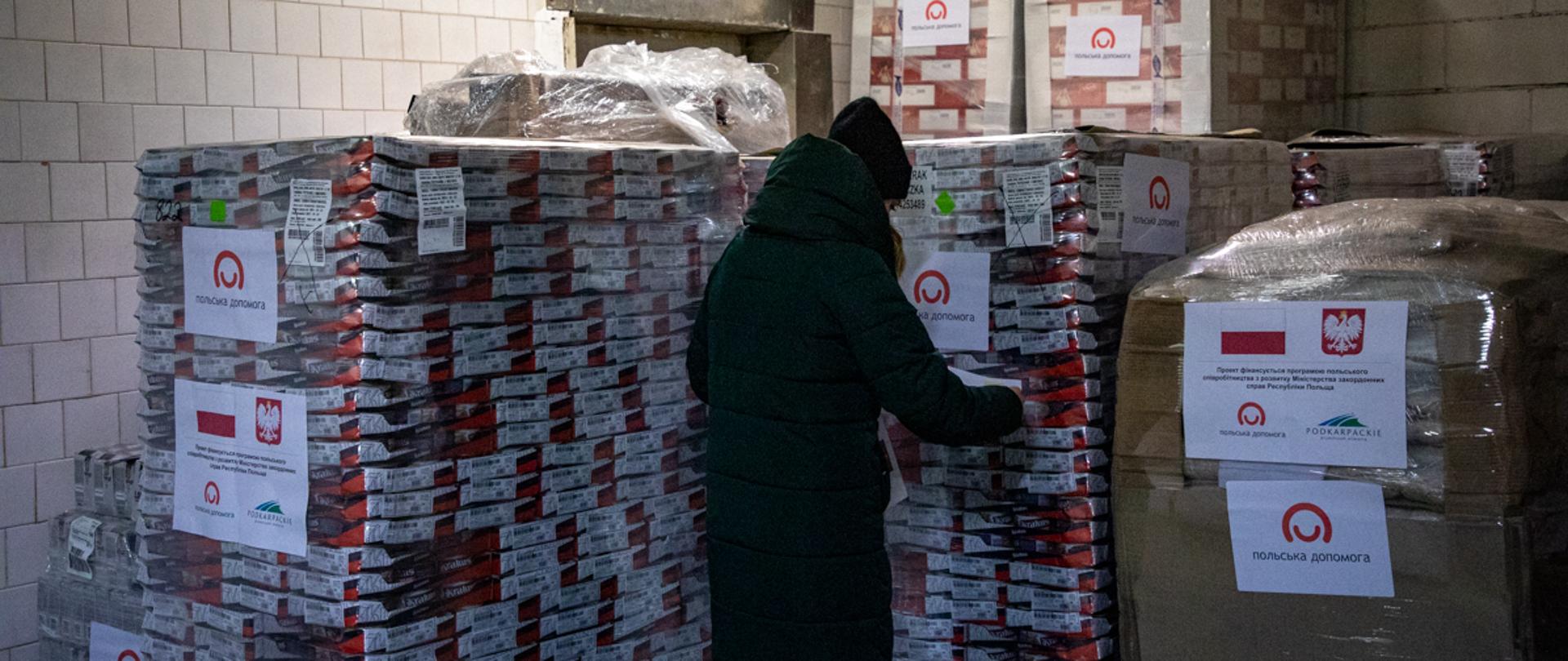 Zdjęcie przedstawia kobietę przyklejającą informację na palety z paczkami żywnościowymi 