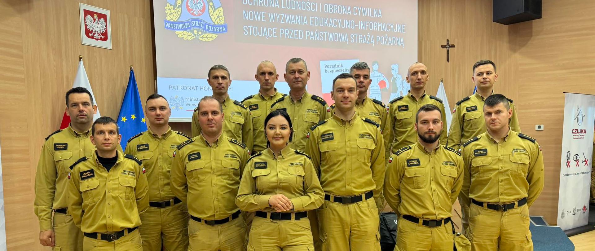 Konferencja pt. „Ochrona ludności i obrona cywilna - nowe wyzwania edukacyjno-informacyjne stojące przed Państwową Strażą Pożarną” - podlascy koordynatorzy ds. prewencji społecznej stoją w żółtych mundurach, stoją w tle hasło Konferencji