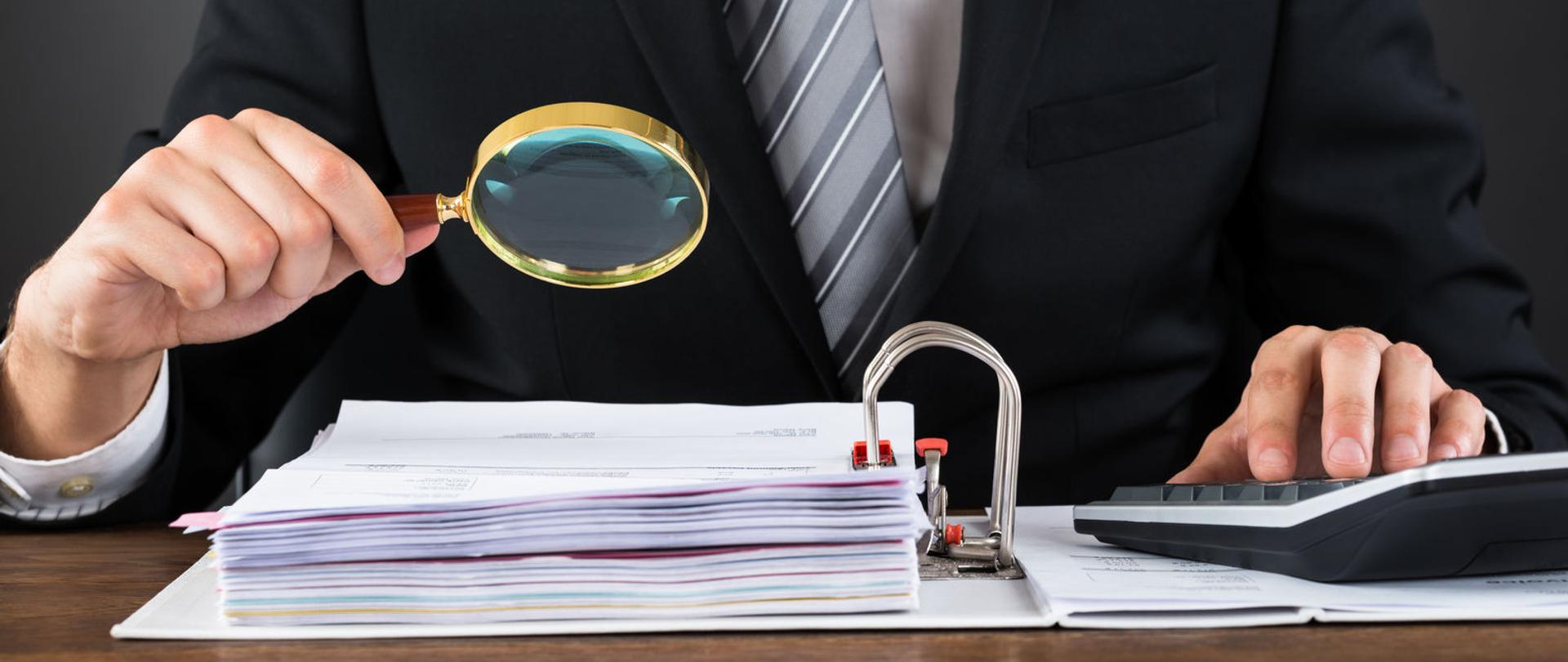 Dłoń trzyma lupę nad segregatorem z dokumentami, druga dłoń obsługuje kalkulator.