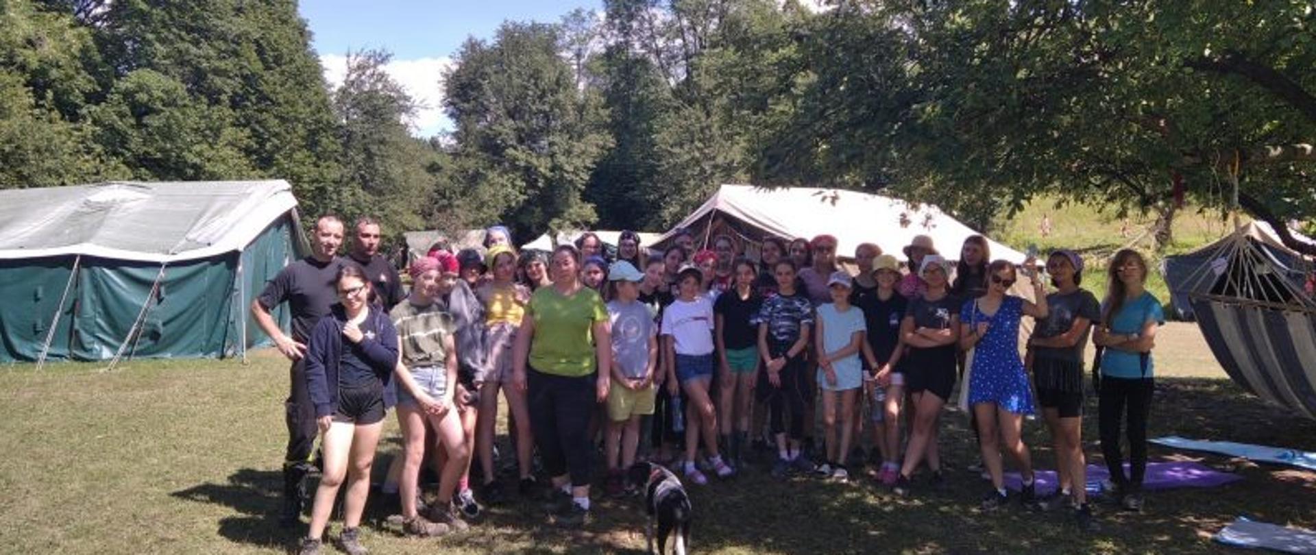 Na zdjęciu dzieci i młodzież uczestnicy obozu wraz z dwoma strażakami z Komendy Powiatowej Państwowej Straży Pożarnej z Jasła pozują do zdjęcia w tle drzewa oraz namioty harcerskie hamak na drzewie, przed cała grupą stoi pies