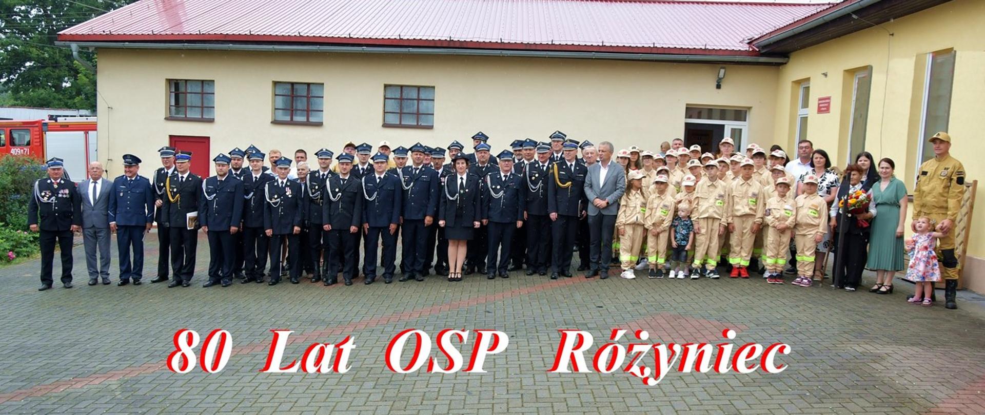 Pamiątkowe zdjęcie grupowe wykonane podczas obchodów 80-lecia Ochotniczej Straży Pożarnej w Różyńcu. Przed budynkiem remizy strażackiej stoją strażacy w mundurach galowych, członkowie Młodzieżowej Drużyny Pożarniczej, zaproszeni goście oraz mieszkańcy. Na zdjęciu widoczny jest napis „80 Lat OSP Różyniec”. W tle znajduje się budynek remizy z czerwonym dachem oraz częściowo widoczny samochód pożarniczy.