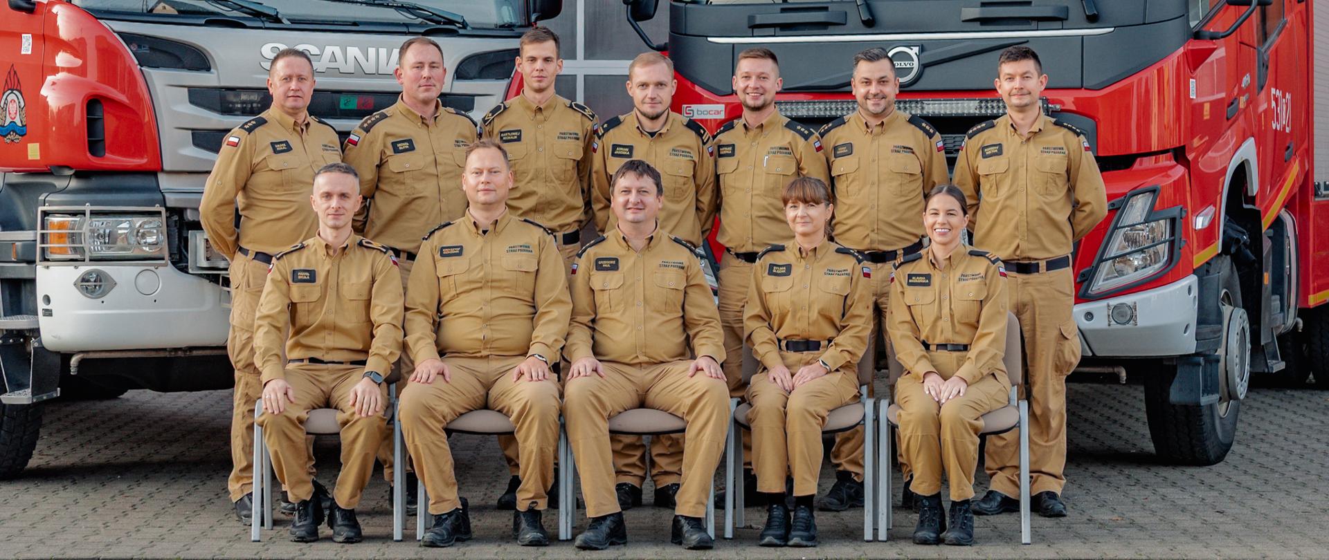Grupa funkcjonariuszy Państwowej Straży Pożarnej w umundurowaniu służbowym pozuje do pamiątkowego zdjęcia na tle dwóch czerwonych samochodów ratowniczo-gaśniczych. W pierwszym rzędzie siedzi pięć osób, a w drugim stoi dziewięć osób. Wszyscy są wyprostowani, uśmiechnięci i ustawieni symetrycznie między pojazdami. Zdjęcie wykonane na placu przed garażami jednostki.