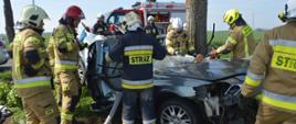 Tragiczny wypadek. Droga z Sarnówki do Łaszczyna. Obok drzewa znajduje sie wrak samochodu osobowego, który brał udział w wypadku. Jest koloru srebrnego. Wokół auta strażacy prowadzący akcje ratowniczą. Jeden z nich pracuje nozycami hydraulicznymi.W tle znajduje się wóz strażacki.