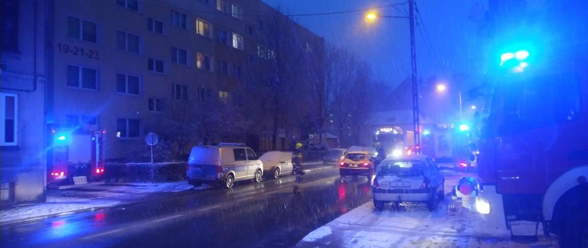 Obraz przedstawia pojazdy pożarnicze zaparkowane przy ul. Starojaworskiej. Pojazdy z włączonymi światłami alarmowymi niebieskimi. Pora nocna. Opady deszczu i śniegu.