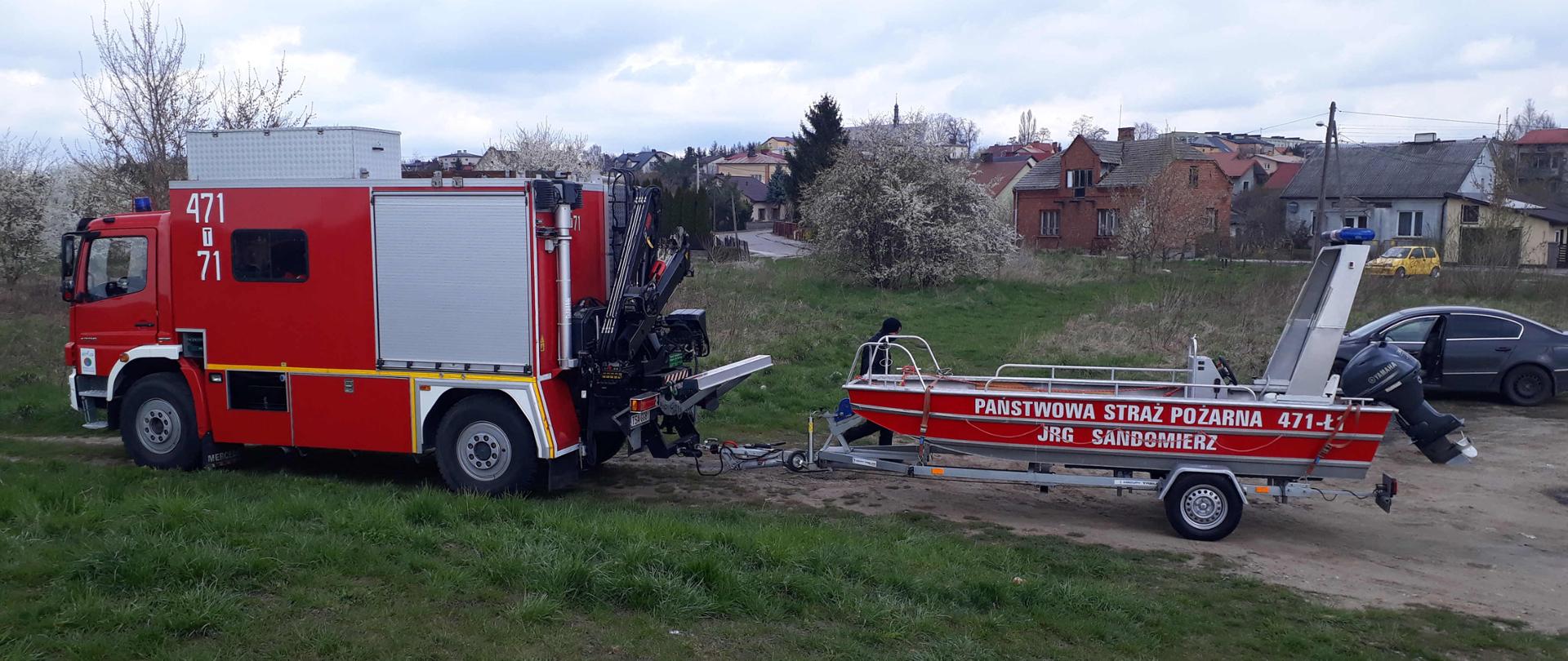 Zdjęcie zrobione na terenie zielonym, w tle zabudowania. W centralnej części zdjęcia znajduje się duży, ciężarowy, czerwony samochód strażacki, który służy nurkom do przemieszczania się. Z tyłu na samochodzie zamontowany jest nieduży dźwig. Do samochodu przypiętą jest przyczepa, na której leży strażacka łódź z silnikiem. Za łodzią widać strażaka niosącego sprzęt do nurkowania.