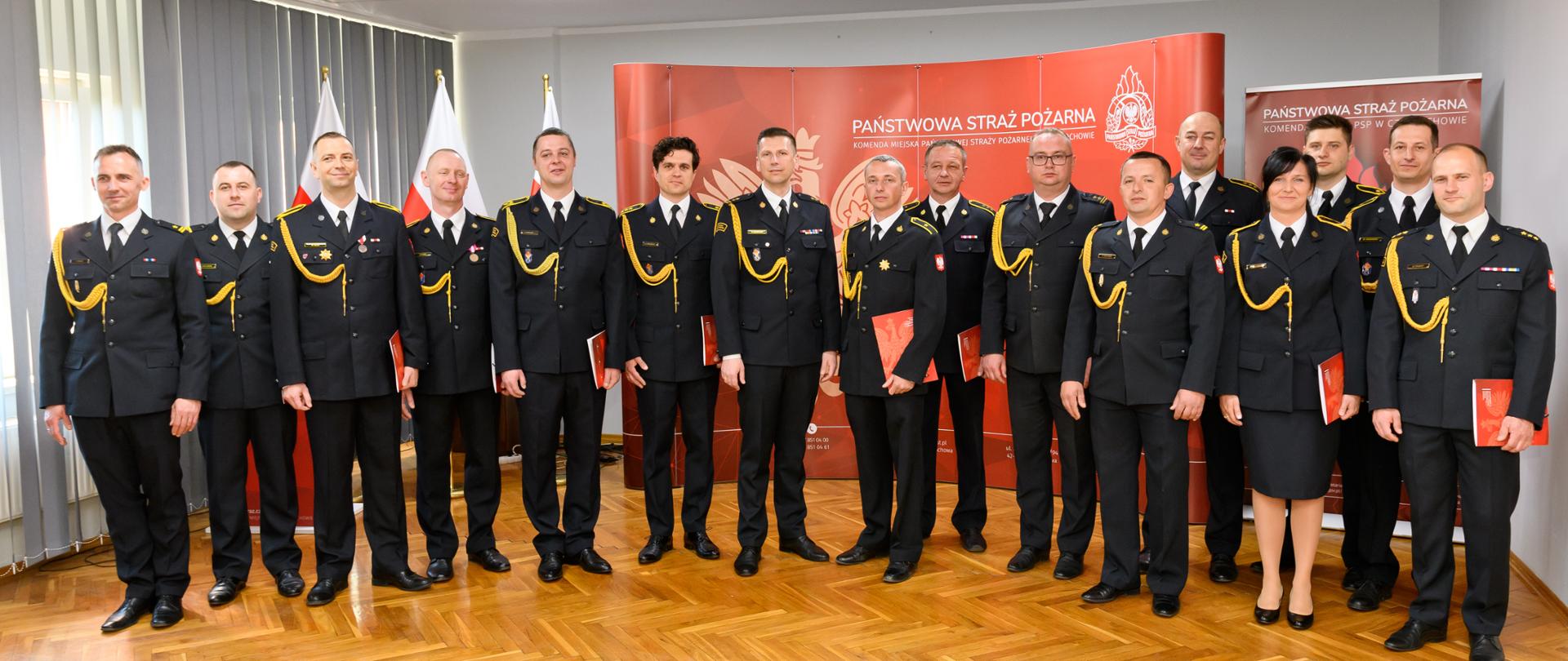 Na zdjęciu grupa funkcjonariuszy Państwowej Straży Pożarnej W mundurach galowych ustawiona do pamiątkowego zdjęcia, niektórzy z nich w rękach trzymają teczki z dokumentami. W tle widoczna jest ścianka z napisem Państwowa Straż Pożarna oraz flagi państwowe.