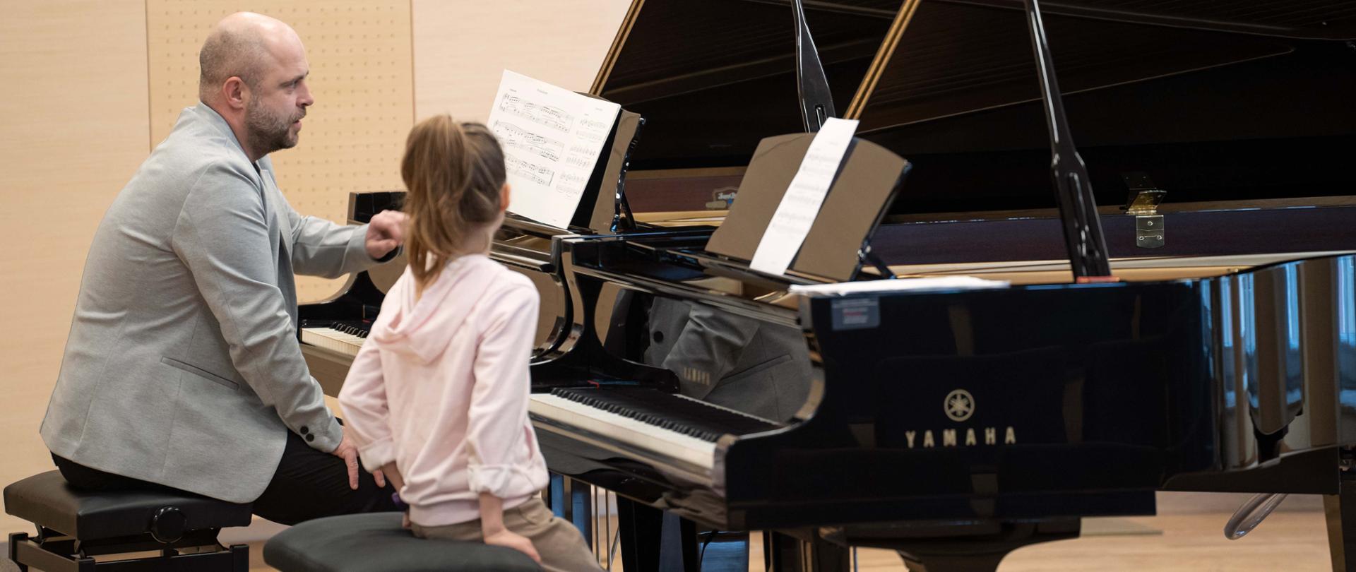zdjęcie przedstawia prowadzącego warsztaty siedzącego przy drugim fortepianie głębiej na scenie sali koncertowej omawiającego tekst nutowy utworu na pulpicie, na pierwszym planie uczennica siedzi słuchając przy pierwszym fortepianie