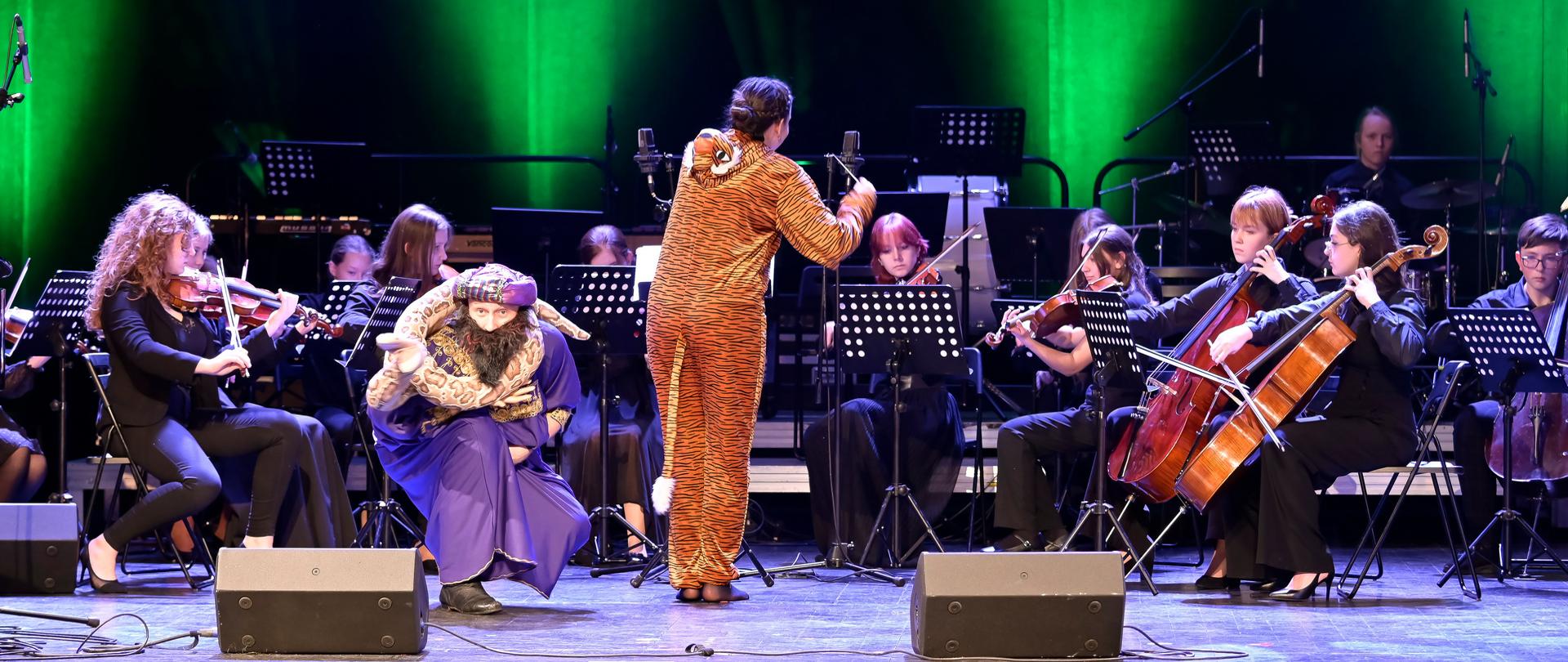 Kobieta dyryguje przebrana w strój tygrysa, obok po lewej stronie kuca mężczyzna w płaszczu trzymający meteriałowego węża, za nimi gra orkiestra.