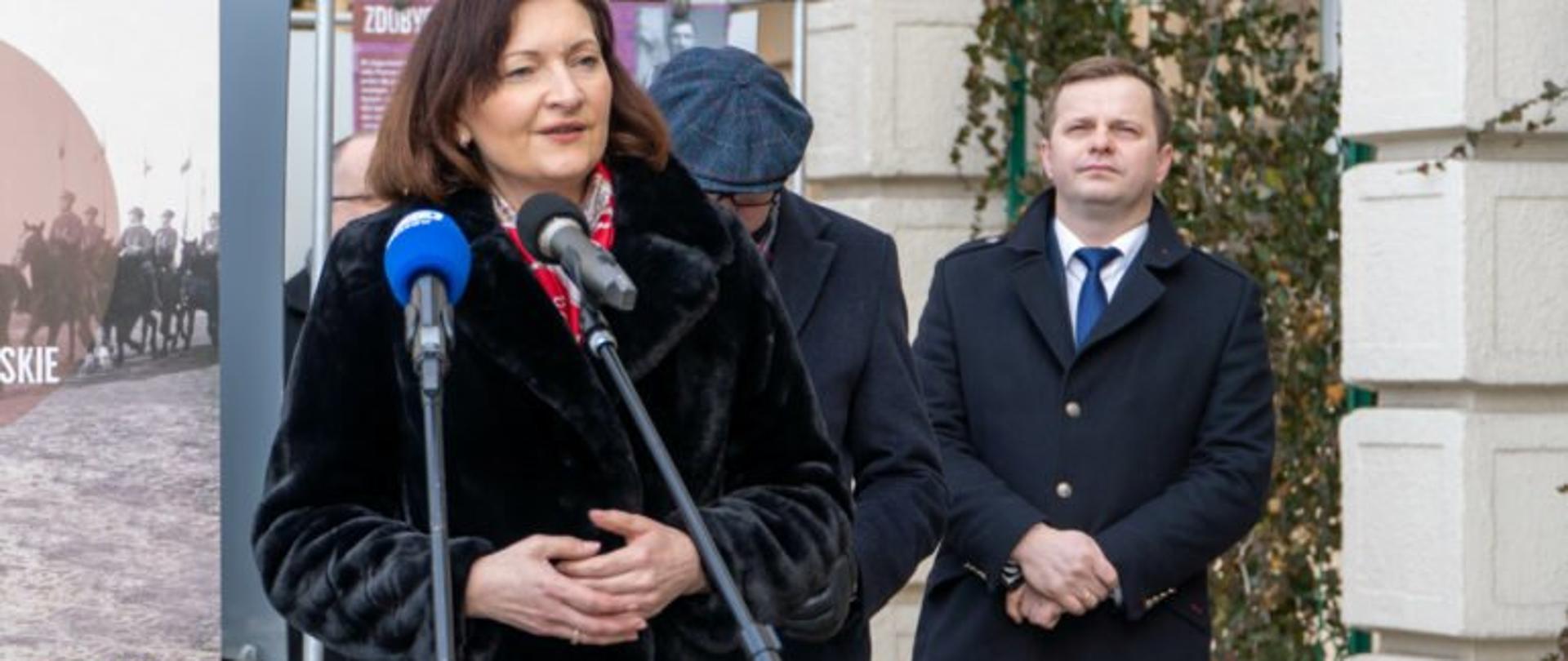 Wojewoda podkarpacki Ewa Leniart zabiera głos podczas uroczystego otwarcia wystawy „Powstanie Wielkopolskie 1918–1919”. W tle fragmenty ekspozycji oraz uczestnicy konferencji prasowej. 