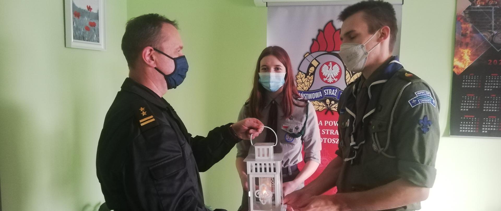 Zdjęcie przedstawia harcerzy z ZHP Krotoszyn, którzy wręczają zastępcy komendanta powiatowego Państwowej straży Pożarnej w Krotoszynie lampion z Betlejemskim Światełkiem Pokoju.