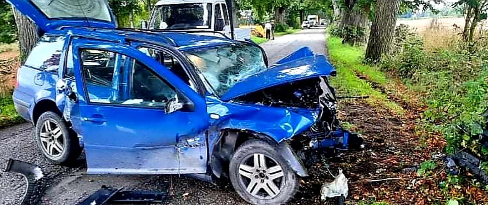 Zdjęcie przedstawia rozbite niebieskie auto na drodze. W tle widać inne pojazdy i drogę.